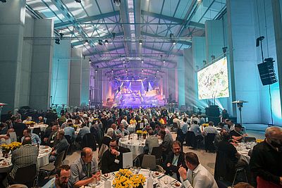 Galadinner findet in einer Festhalle statt. Auf dem Tisch befinden sich gelbe Blumen. Ganz vorne befindet sich ein Kieshaufen, der als Bühne genutzt wird.